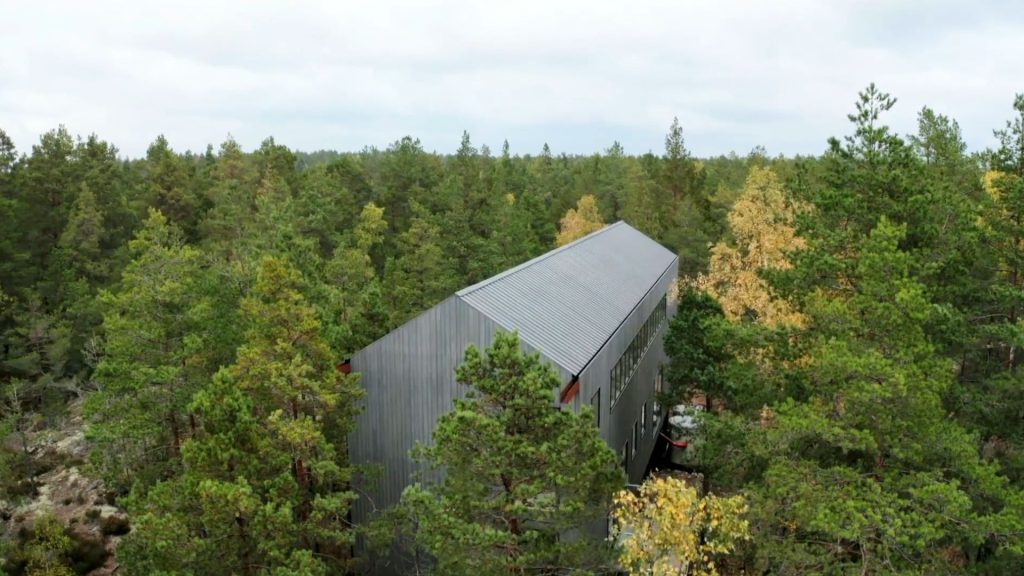 Stiligt - En modern, grå byggnad med plåttak är omgiven av tät skog med grönt och gult bladverk under en molnig himmel.