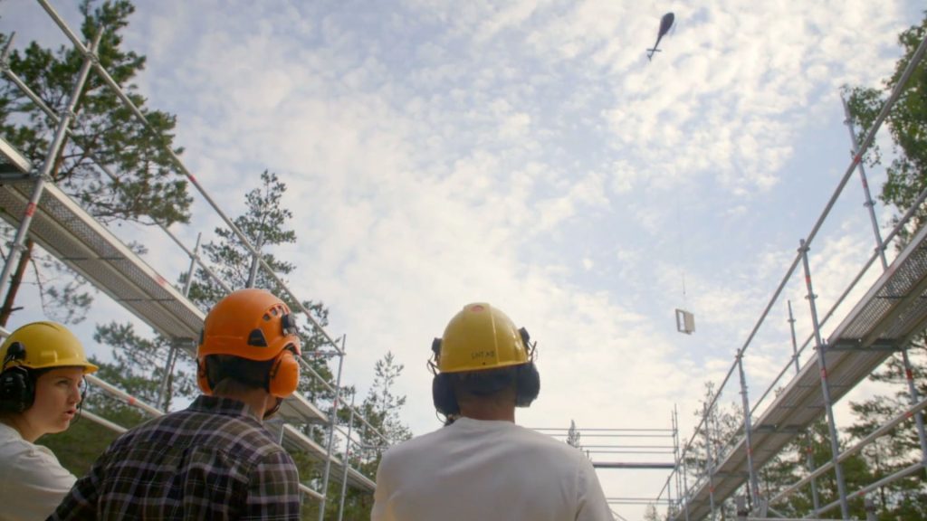 Stiligt - Tre byggnadsarbetare med hjälm och hörselskydd tittar upp när en helikopter transporterar byggmaterial ovanför en arbetsplats med byggnadsställningar och träd.