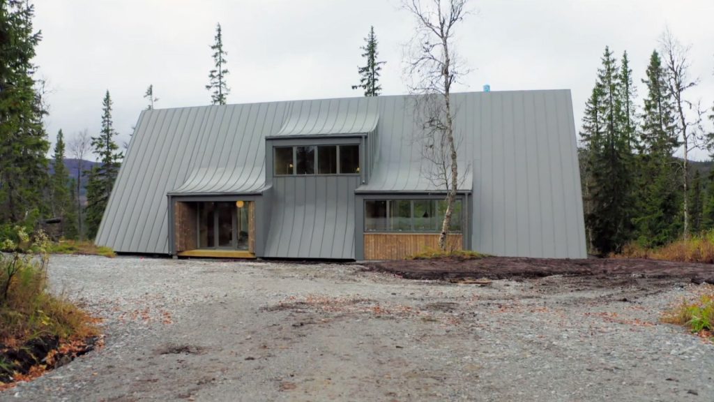 Stiligt - Ett modernt hus med brant plåttak och stora fönster, omgivet av träd och grus, ligger i ett skogsområde under en mulen himmel.