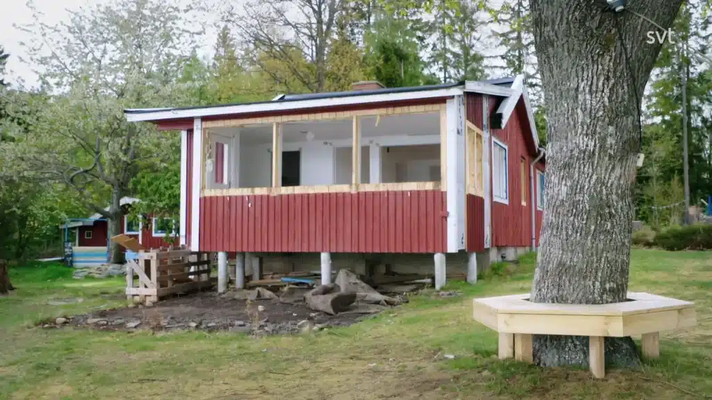 Stiligt - Ett rött trähus på pålar är under renovering, med synlig inramning och inga fönsterrutor; en träbänk omger ett närliggande träd.