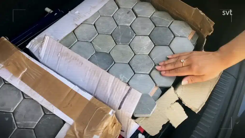Stiligt - En persons hand vilar på en låda fylld med sexkantiga grå plattor, en del förpackningar och kartongkanter synliga runt plattorna.