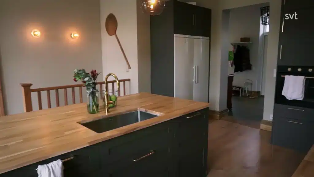 Stiligt - Modernt kök med bänkskiva i trä, mässingsblandare, mörka skåp, väggmonterad träslev och inbyggda vitvaror. Blommor i en vas placeras på ön.