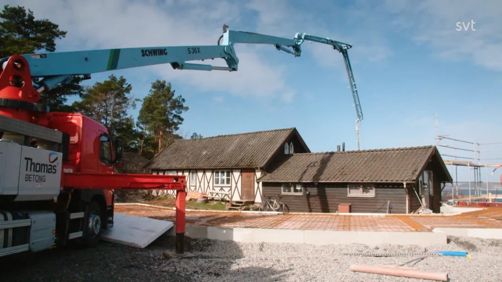 Stiligt - En betongpumpbil häller betong på en byggarbetsplats framför ett trähus med tegeltak en klar dag.