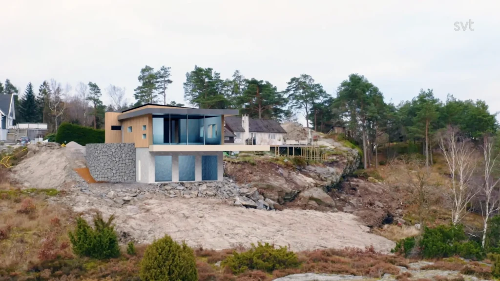Stiligt - Ett modernt hus med stora fönster står på ett stenigt, upphöjt landskap omgivet av träd och andra hus under en molnig himmel.