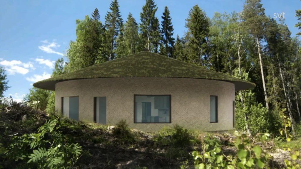 Stiligt - Ett cirkulärt hus med ett grönt, mossbeklätt tak står i ett skogsområde, omgivet av träd och buskar under en klarblå himmel.