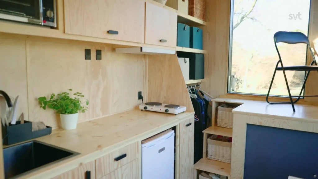 Stiligt - Kompakt kök med ljusa träskåp, ett litet kylskåp, hyllor med förvaringskorgar, en enda planta och ett bord vid ett stort fönster med en hopfällbar stol.