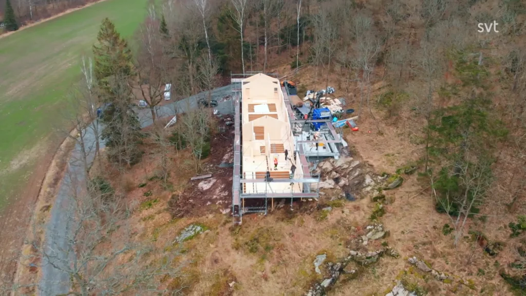 Stiligt - Flygfoto av ett hus under uppbyggnad i ett skogsområde, med byggmaterial och utrustning utspridda runt platsen.