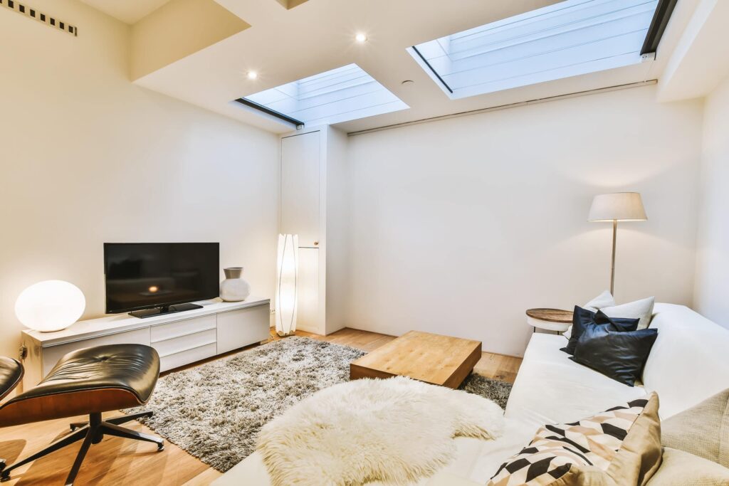 Stiligt - Modernt vardagsrum med en vit sektionssoffa, soffbord i trä, TV på en låg konsol, matta, fåtölj, golvlampa och takfönster som ger naturligt ljus.