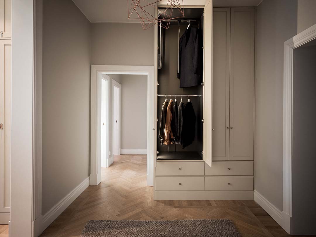 Stiligt - Modern hall med inbyggd beige garderob, några kläder hängande inuti, ljust fiskbensgolv i trä, neutrala väggar och en geometrisk taklampa.