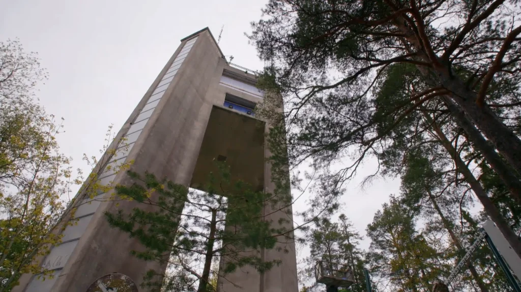Stiligt - Högt utsiktstorn i betong med en öppen mittdel, omgiven av träd, sett från en låg vinkel mot en molnig himmel.