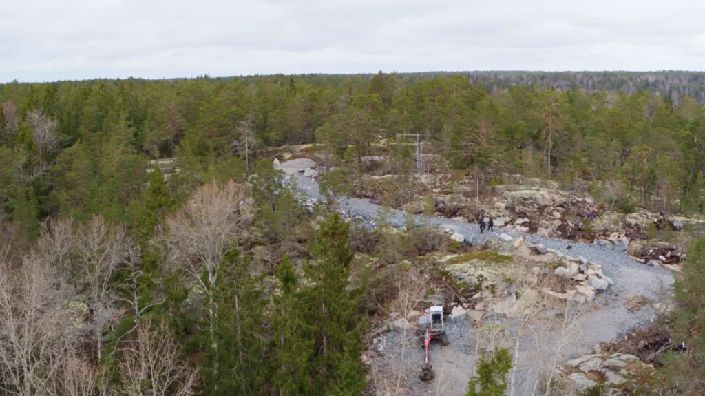 Stiligt - En grusväg skär genom ett skogsområde med träd och gles vegetation under en molnig himmel; anläggningsutrustning är synlig på vägen.