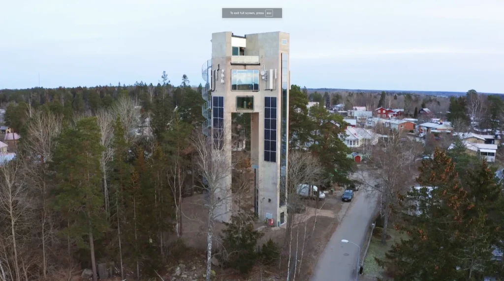 Stiligt - En hög, smal betongbyggnad med stora fönster och solpaneler står bland träd nära en väg i ett bostadsområde.