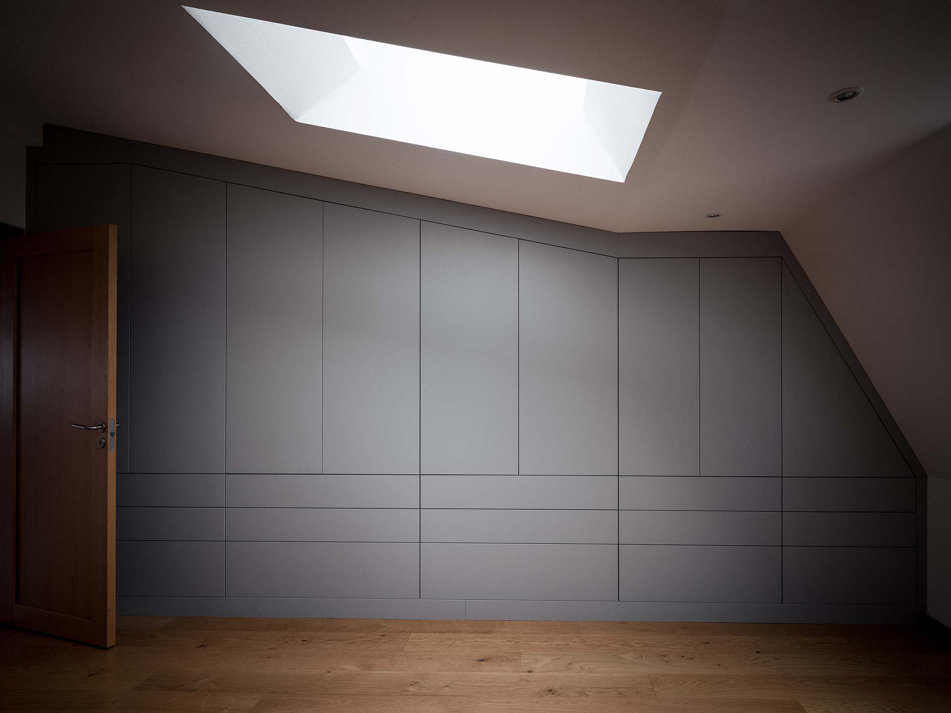 Stiligt - En inbyggd grå garderob passar mot snedtak under ett stort takfönster, med trägolv och öppen trädörr till vänster.