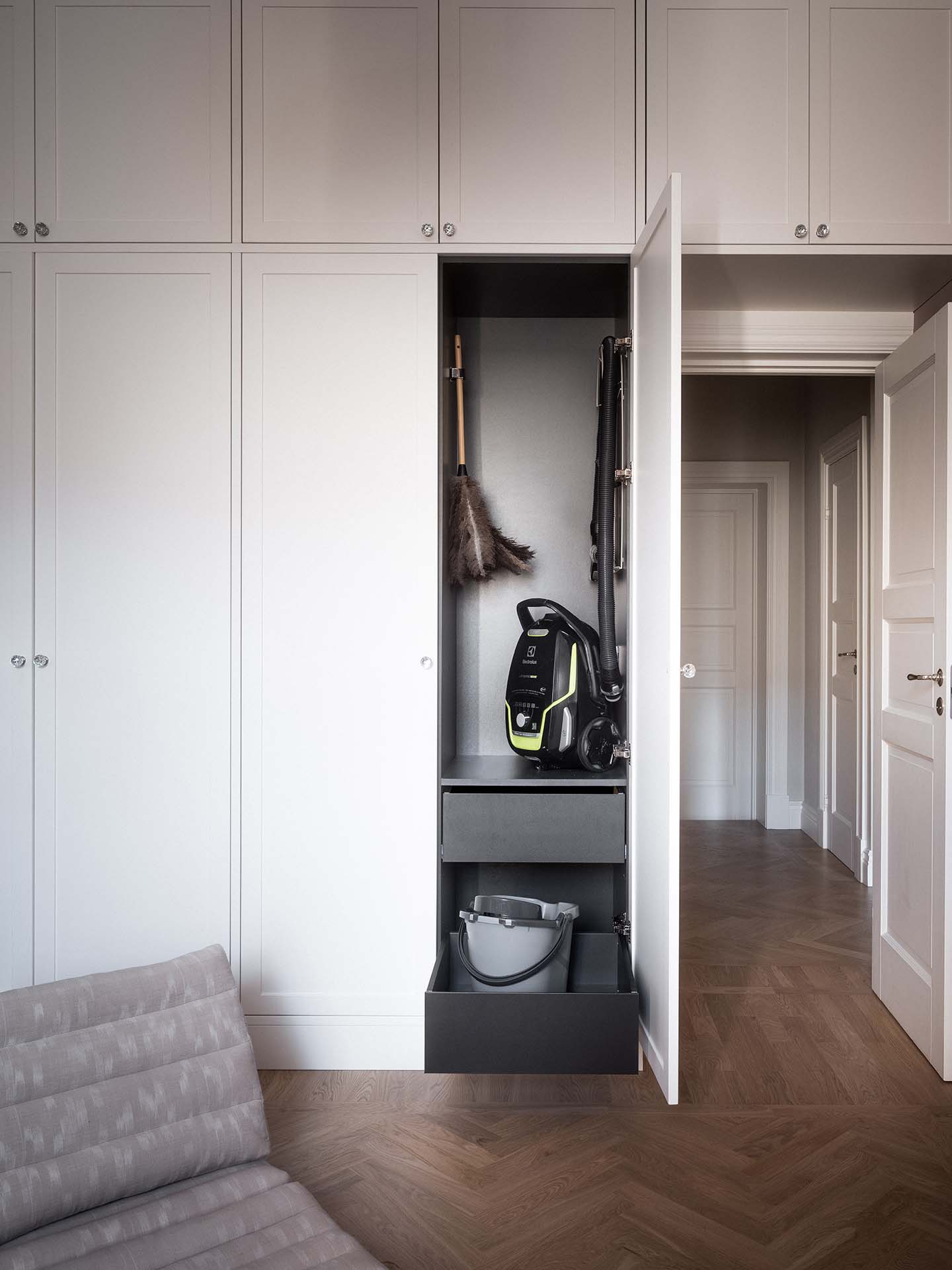 Stiligt - Ett högt vitt skåp med en dörr öppen visar en dammsugare, en dammduk, ett mopphandtag och en hink inuti. Rummet har trägolv och vita väggar.