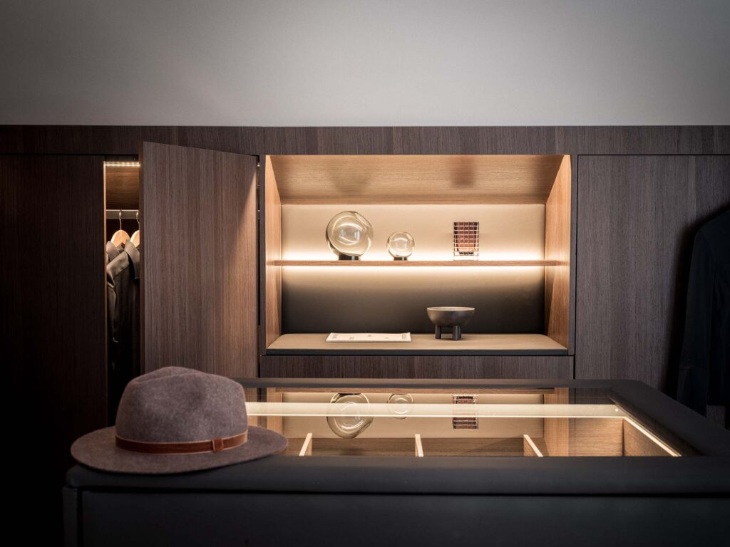 Stiligt - En modern garderob i mörkt trä med öppna hyllor visar dekorativa föremål; en fedora vilar på en glasmonterad monter i förgrunden.