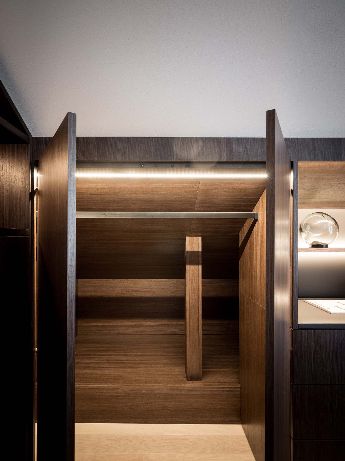 Stiligt - Öppen träskåp med mörka träpaneler, inbyggda hyllor och en horisontell hängstång, upplyst av takbelysning.
