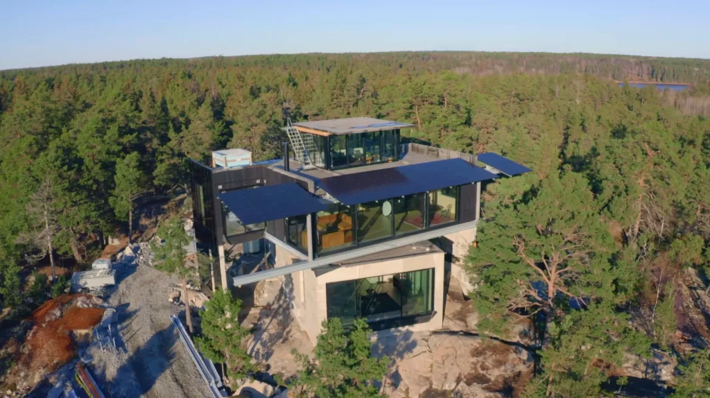 Stiligt - Ett modernt hus i glas och betong med solpaneler är byggt på en klippig sluttning och omgivet av tät skog under en klarblå himmel.