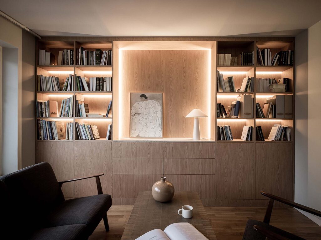 Stiligt - En modern inbyggd bokhylla i trä med integrerad belysning, fylld med böcker och dekor, med konst och en lampa i en central alkov; ett bord med en vas, kopp och öppen bok i förgrunden.