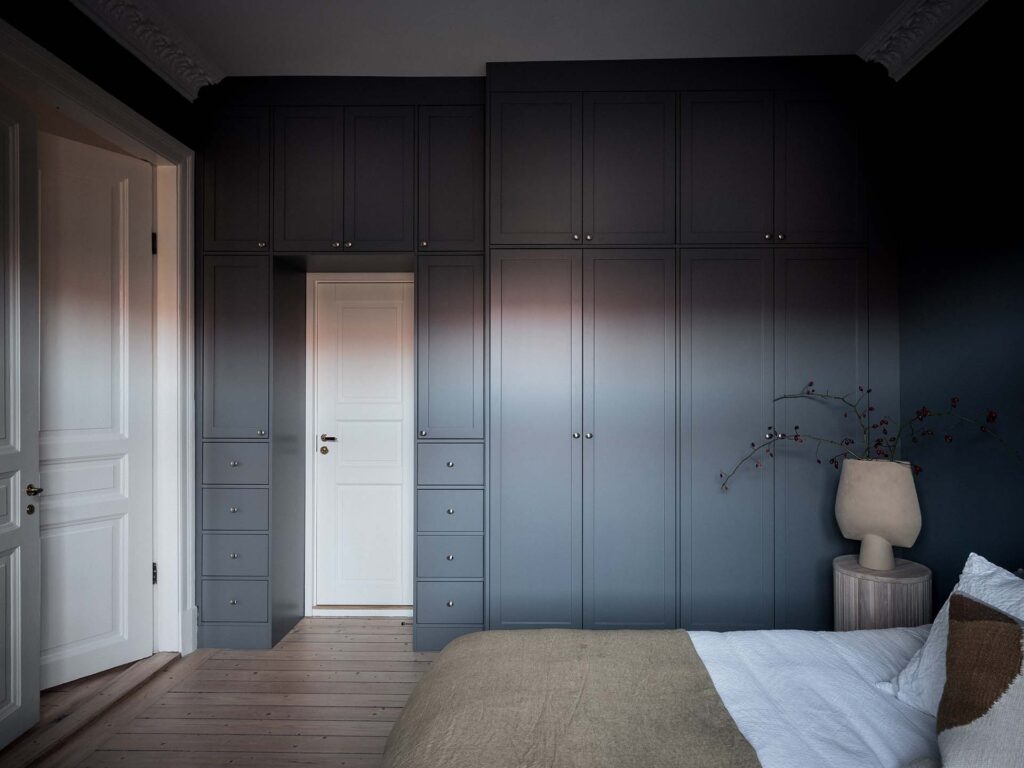 Stiligt - Sovrum med inbyggda grå garderober, stängd vit dörr, ljust trägolv och en säng med vita sängkläder och beige plagg, bredvid ett sidobord med lampa och vas.
