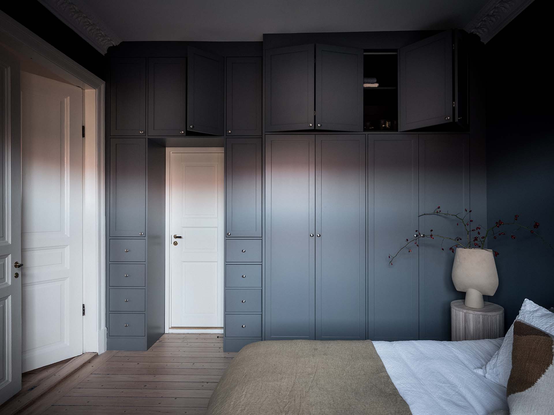 Stiligt - Sovrum med inbyggda grå garderober, stängd vit dörr, ljust trägolv och en säng med vita sängkläder och beige plagg, bredvid ett sidobord med lampa och vas.