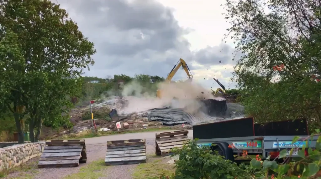 Stiligt - En grävmaskin river en struktur och skapar ett moln av damm; träpallar och en trailer är i förgrunden, med träd och molnig himmel i bakgrunden.