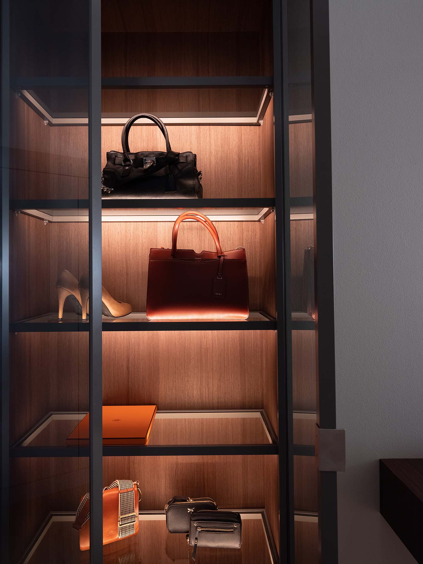 Stiligt - Ett vitrinskåp med trähyllor visar handväskor, högklackade skor och en liten låda, allt upplyst av inbyggd belysning.