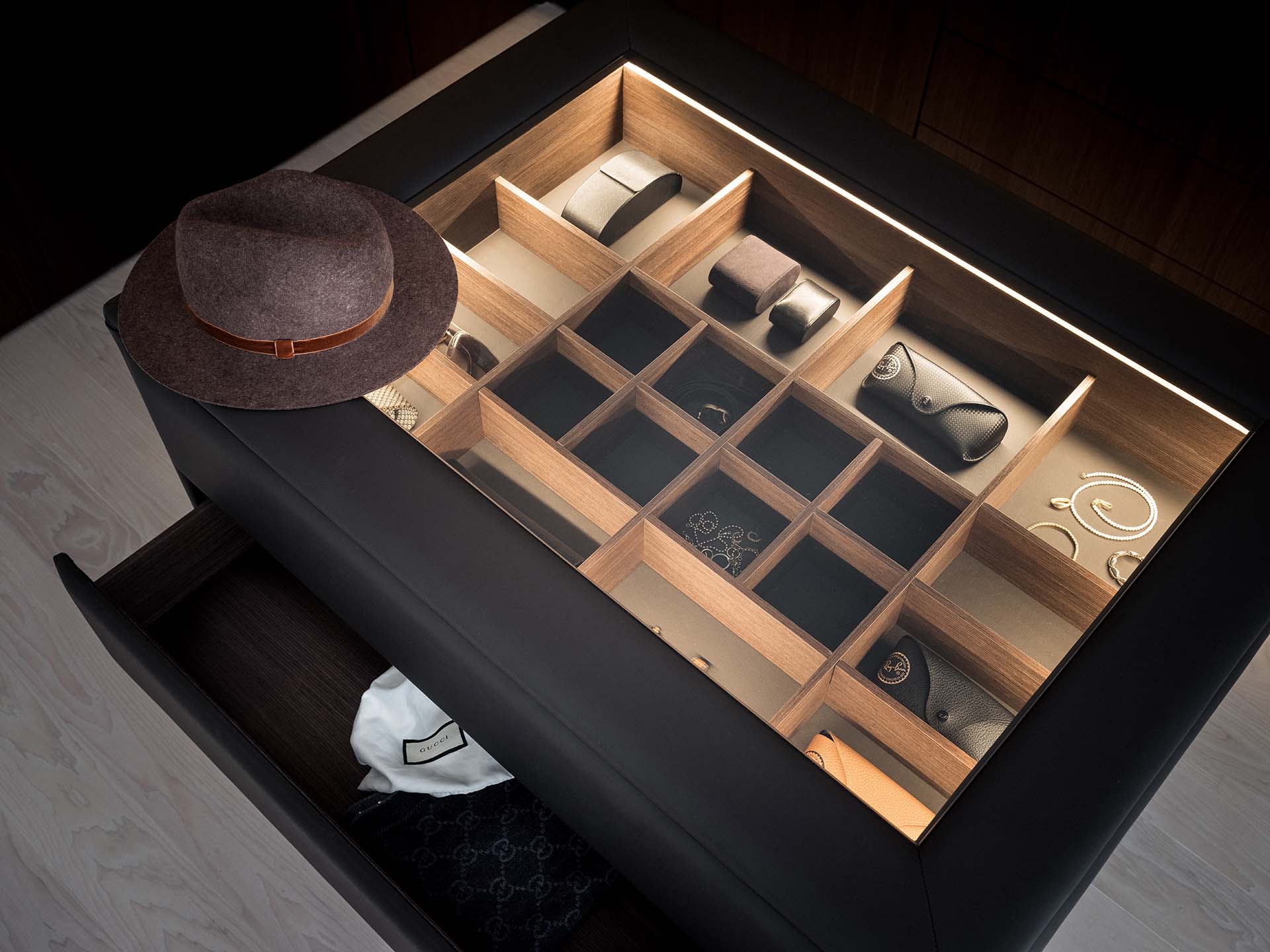Stiligt - En låda med en glasskiva visar organiserade fack som innehåller klockor, solglasögon och smycken; en brun hatt vilar på lådans hörn.