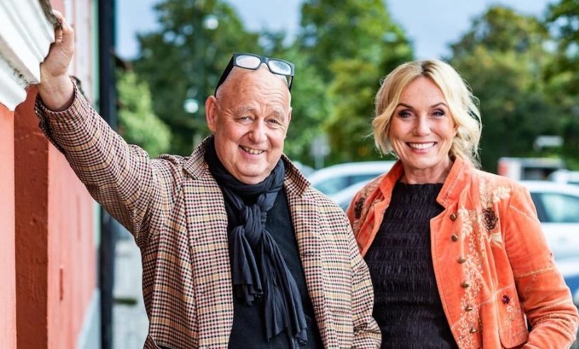 Stiligt - Två leende vuxna, en man och en kvinna, står utomhus framför en byggnad, klädda i mönstrade jackor och tittar på kameran. Träd och parkerade bilar är i bakgrunden.