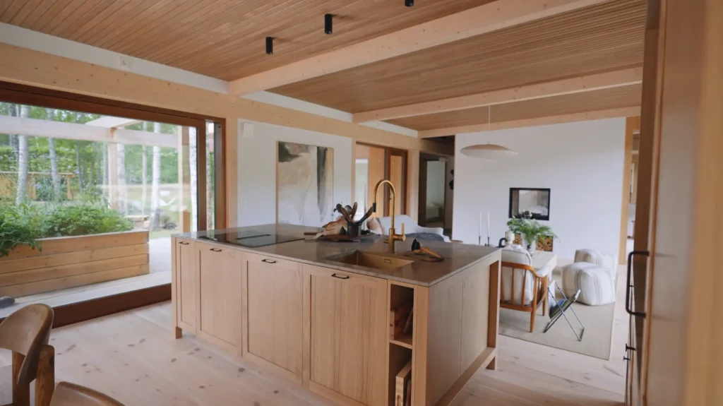 Stiligt - Modernt kök i öppen planlösning med en central träö, inbyggd diskho och stora fönster med utsikt över en uteplats med växter. Ljusa trätoner och minimalistisk inredning i hela utrymmet.