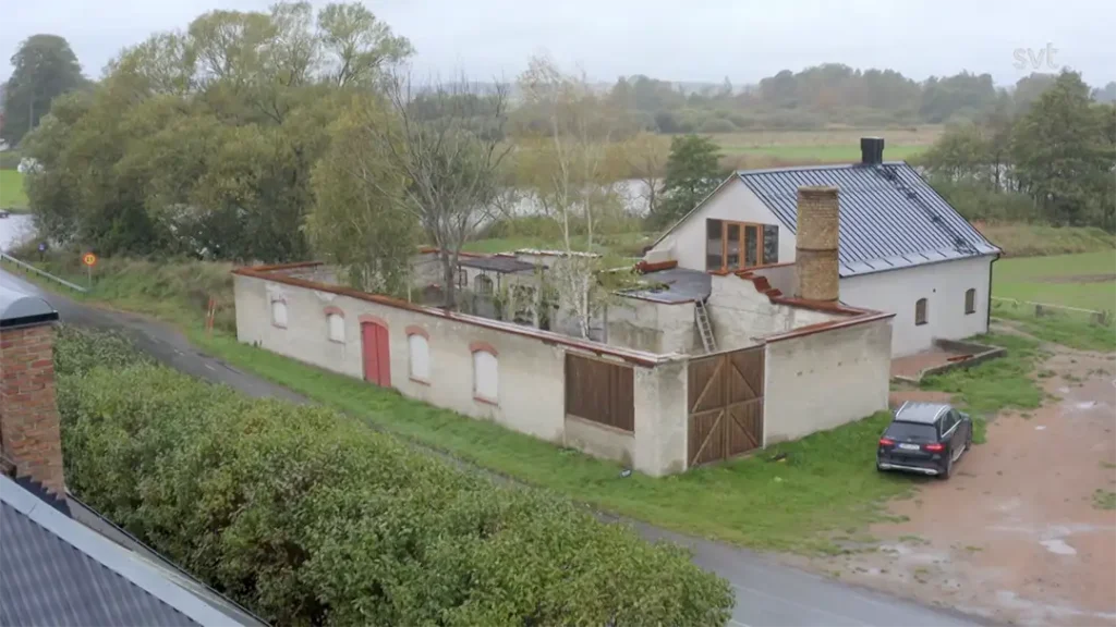 Stiligt - En vit byggnad med plåttak står bredvid ruinerna av en gammal tegelkonstruktion; en bil står parkerad i närheten på en landsväg omgiven av grönska.