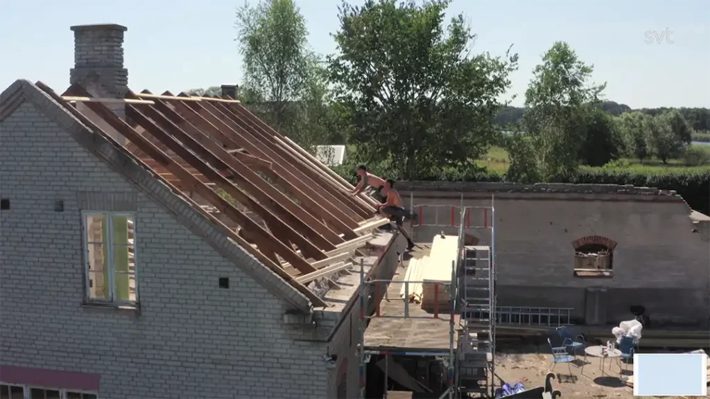 Stiligt - Två personer arbetar på träramen av ett delvis konstruerat tak ovanpå en tegelbyggnad, med ställningar och byggmaterial nedan. Träd och fält syns i bakgrunden.