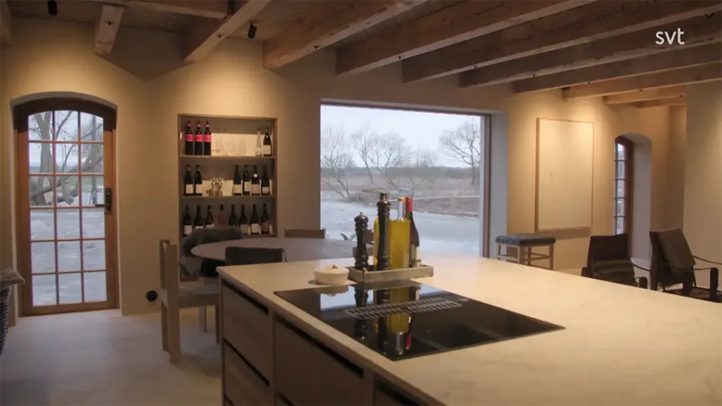 Stiligt - Modernt kök med synliga träbjälkar, en marmorö, spis, vinställ, matbord och stora fönster med utsikt över ett vinterlandskap.