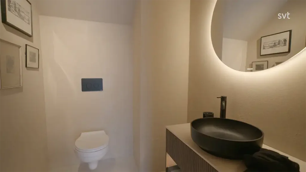 Stiligt - Minimalistiskt badrum med en väggmonterad toalett, ett handfat i svart kärl på en trähandfat, en rund bakgrundsbelyst spegel och inramade konstverk på krämfärgade väggar.