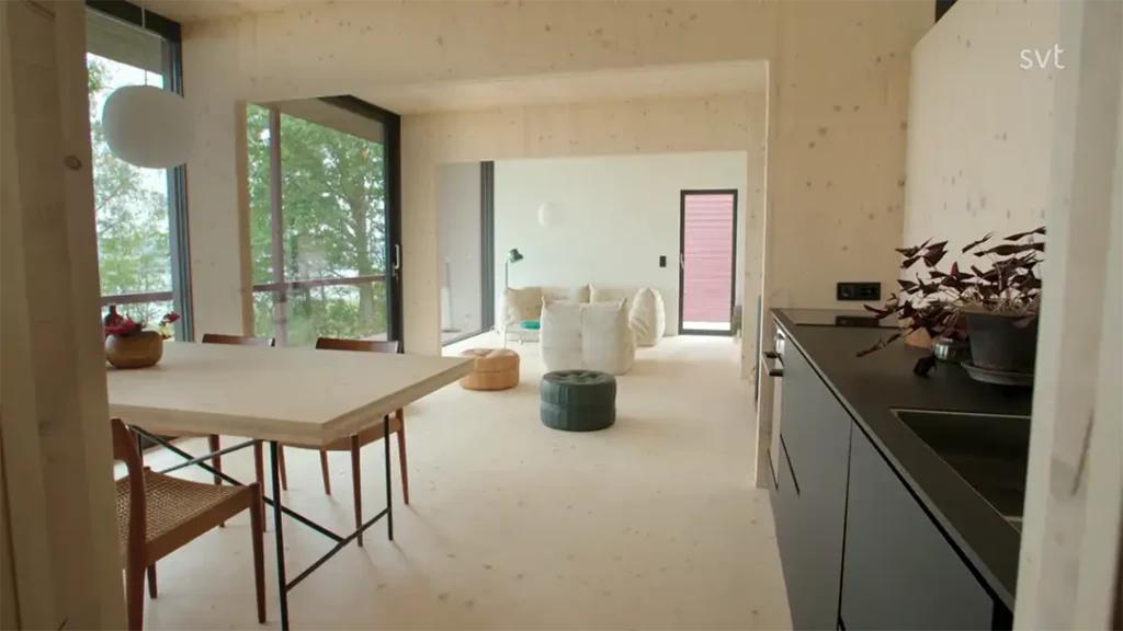 Stiligt - Modernt vardagsrum i öppen planlösning med ljusa trägolv och väggar, ett matbord med stolar, ett litet kök och en sittgrupp med stora fönster med utsikt över träden.