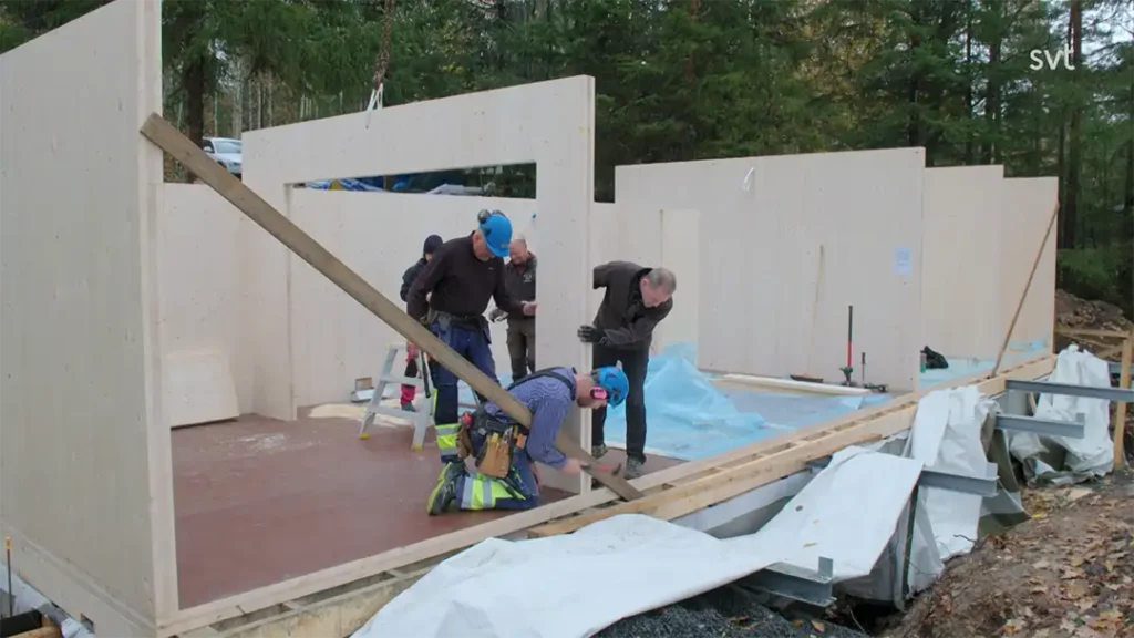 Stiligt - Flera personer monterar träväggpaneler för en byggnad under uppförande på en skogbevuxen plats, med verktyg och material utspridda i närheten.