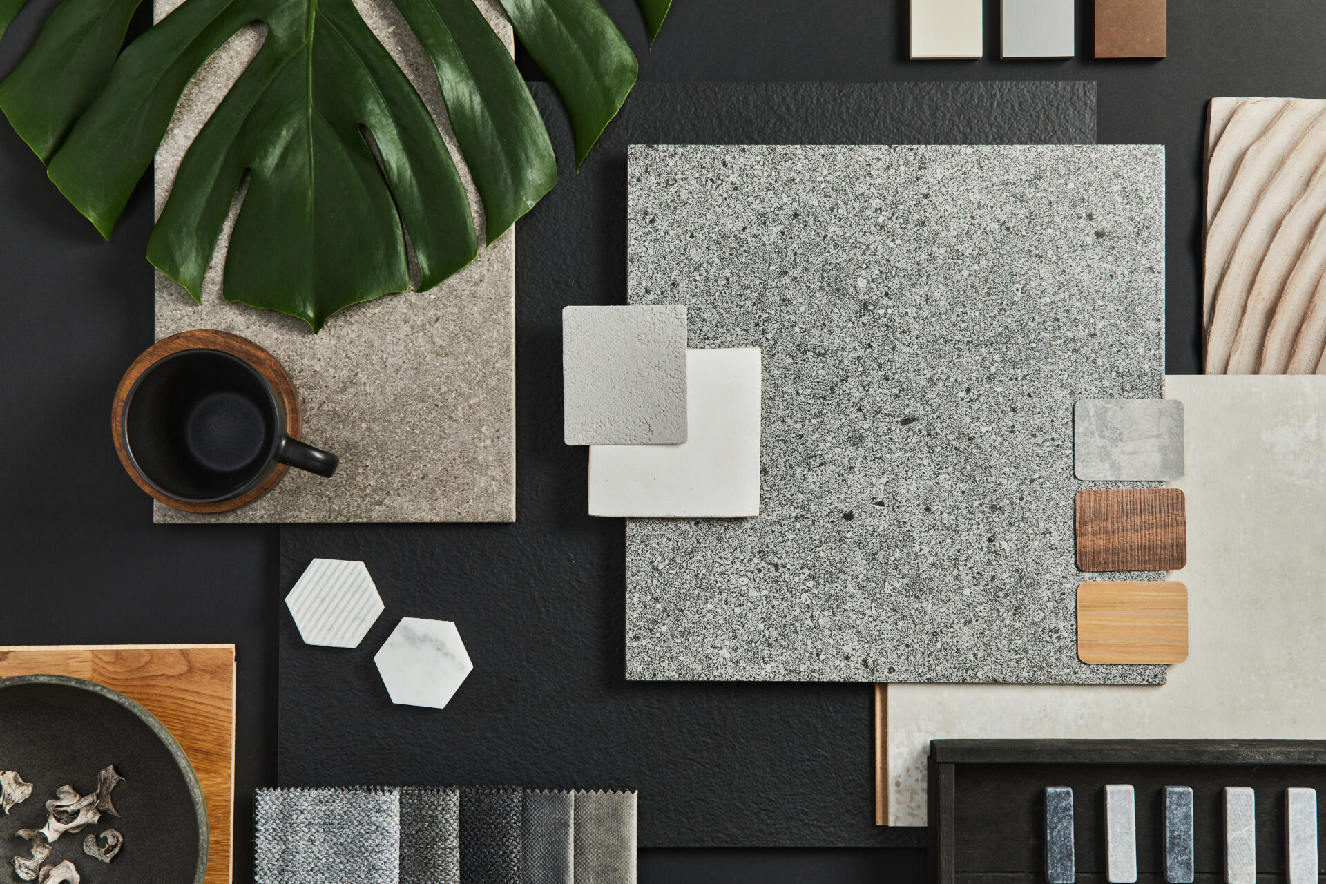 Stiligt - Blandade materialprover, kakel, tygprover, en kopp kaffe och ett stort grönt blad arrangerat på en mörk yta.