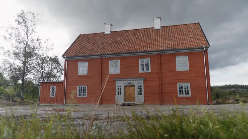 Stiligt - Ett stort rött trähus i två våningar med tegeltak och vita detaljer står mot en molnig himmel, sett från marknivå.