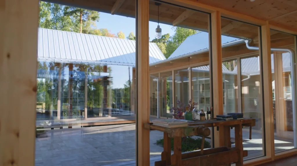 Stiligt - Utsikt genom stora glasfönster som visar en modern innergård med träbjälkar, plåttak och uteservering, omgiven av grönska.