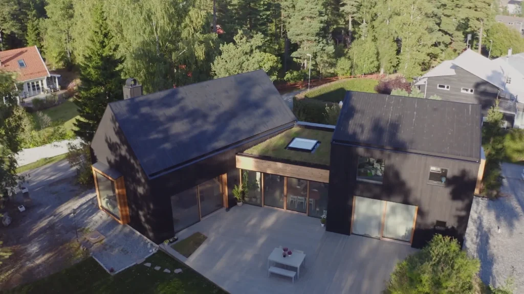 Stiligt - Ett modernt svart hus med två sammanhängande sektioner, stora fönster och en rymlig uteplats, omgiven av träd och grönska i ett skogsområde.