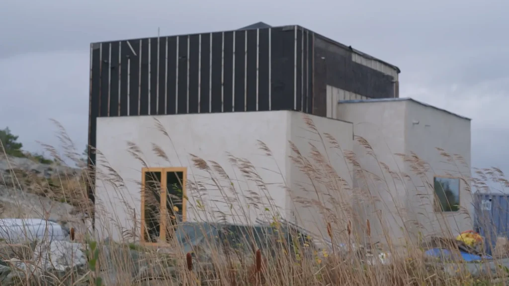 Stiligt - Ett modernt, boxigt hus med vita och mörka fasader ligger bakom höga, torra gräs under en mulen himmel.