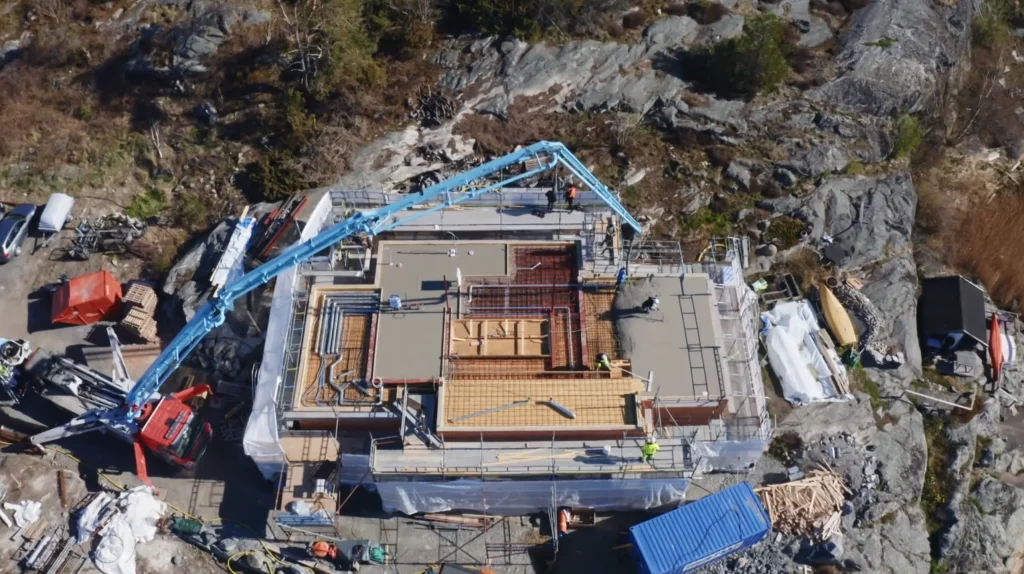 Stiligt - Flygfoto över en byggarbetsplats med arbetare, byggnadsställningar, en betongpumpbil och byggmaterial omgiven av stenig terräng och träd.