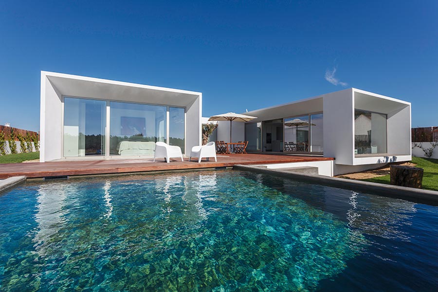 Stiligt - Modernt vitt hus med stora glasfönster, ett trädäck, uteservering och en klarblå pool i förgrunden under en klar himmel.