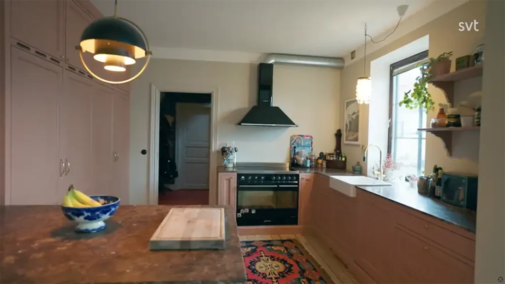 Stiligt - Ett modernt kök med rosa skåp, svart spis och köksfläkt, mönstrad matta och bänkskiva med fruktskål och skärbräda. Naturligt ljus kommer in genom ett fönster.