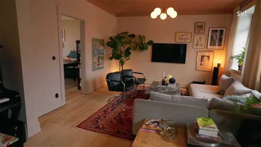 Stiligt - Ett mysigt vardagsrum med en soffa, två stolar, en TV på väggen, inramad konst, en röd matta, krukväxter och varm belysning. En dörröppning leder till ett annat rum.