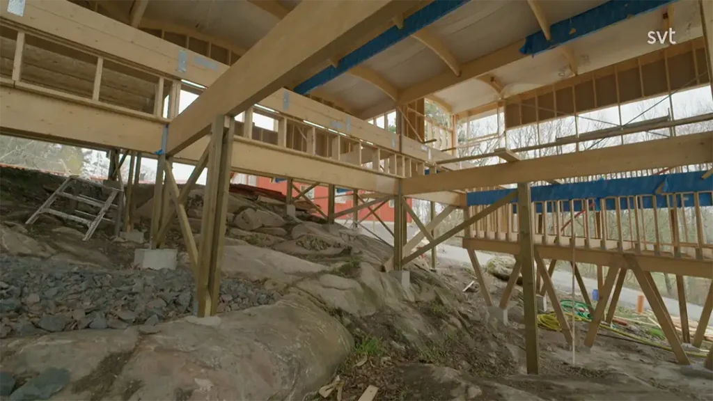 Stiligt - En träbyggnadsram är konstruerad på en stenig, ojämn mark. Utspridda byggmaterial och en stege syns under och runt konstruktionen.