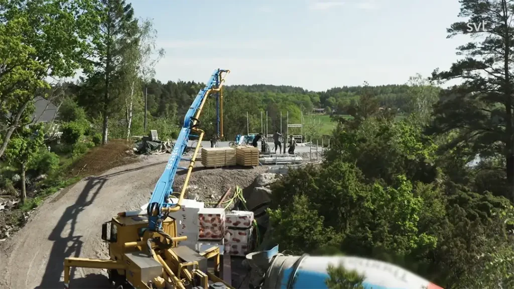 Stiligt - En byggarbetsplats med arbetare, byggmaterial, en kran och en cementbil omgiven av träd och grönska en solig dag.
