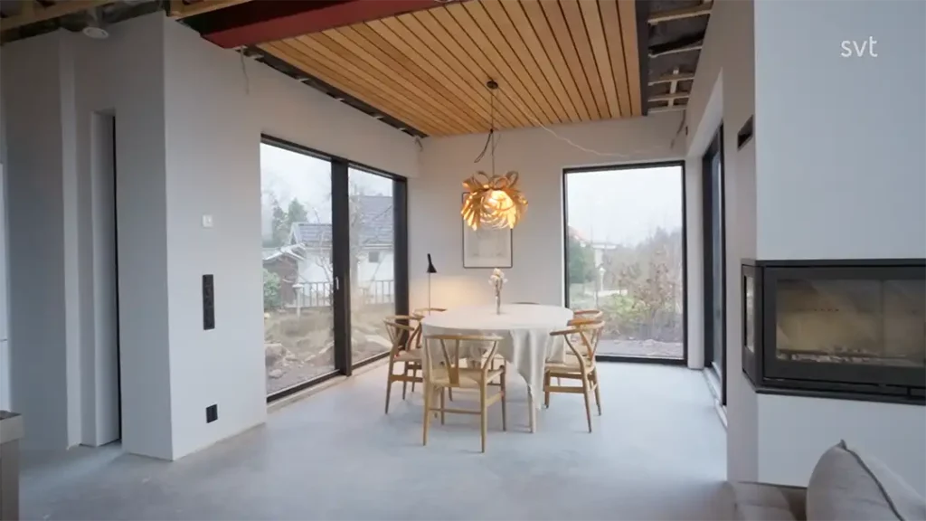 Stiligt - En modern matplats med ett runt bord, fyra trästolar, stora fönster, en taklampa och ett delvis exponerat tak.