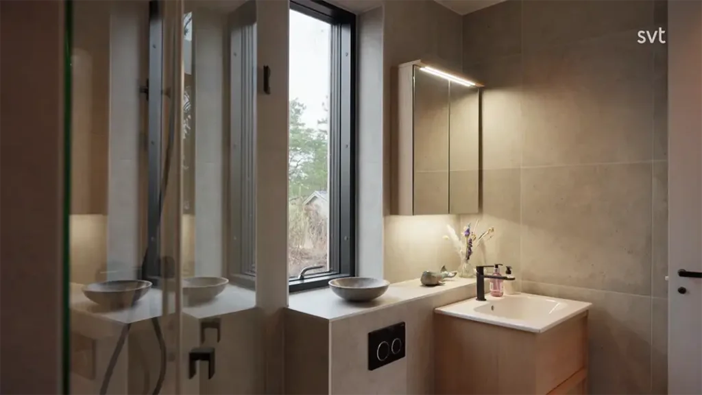 Stiligt - Modernt badrum med stort fönster, väggmonterad toalett, handfat för kärl och upplyst spegel ovanför ett smycke i ljust trä; neutralt tonade kakel genomgående.