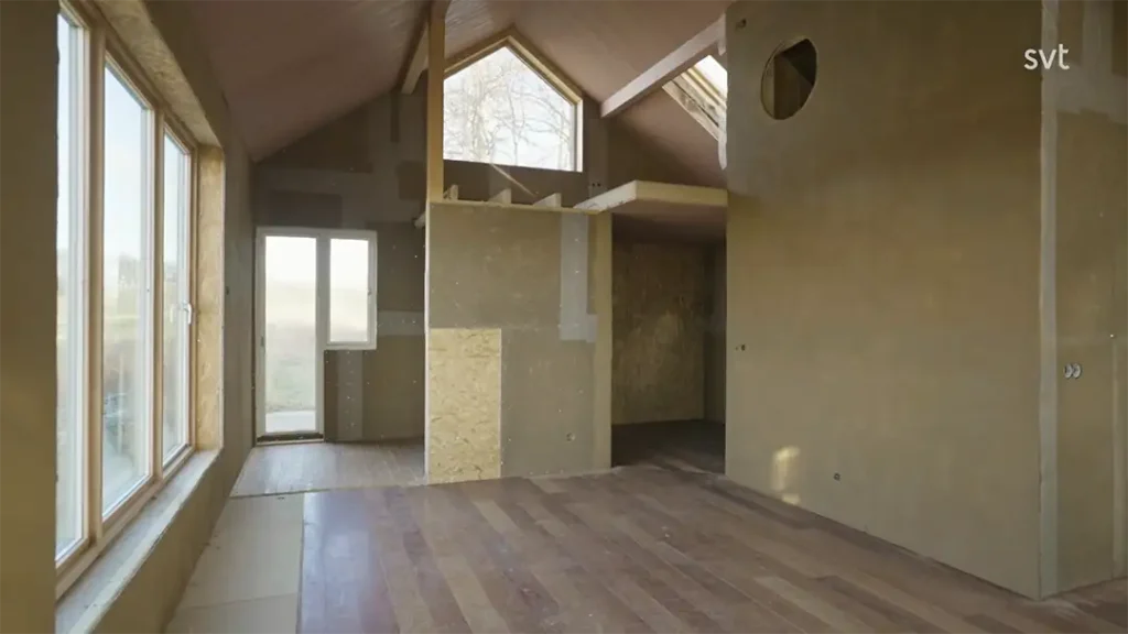 Stiligt - Interiör av ett oavslutat hus med synliga plywoodväggar, stora fönster, ett loftområde och synliga byggmaterial.