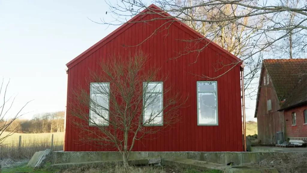 Stiligt - Ett rött trähus med tre höga rektangulära fönster, ett lövlöst träd framför och ett gräsbevuxet fält i bakgrunden.